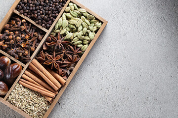 Assortment of whole Indian spices displayed in a wooden box with compartments, featuring cinnamon, star anise, cloves, and cardamom on a textured gray surface with copy space