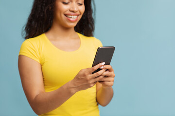 Fototapeta premium Chatting with friends. Happy black woman using smartphone texting sms message or scrolling social networks feed, standing over blue studio background, selective focus