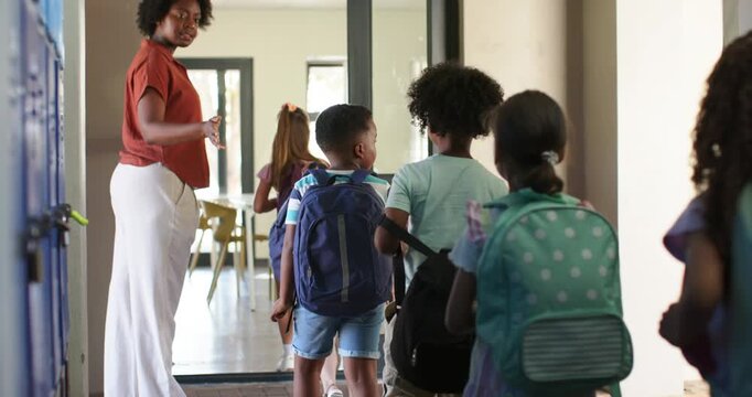 African American teacher gesturing, guiding children down school hall into doorway with backpacks