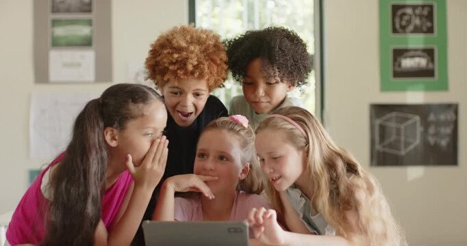 Diverse children gathering around tablet on table in classroom, right girl tapping, sharing focus