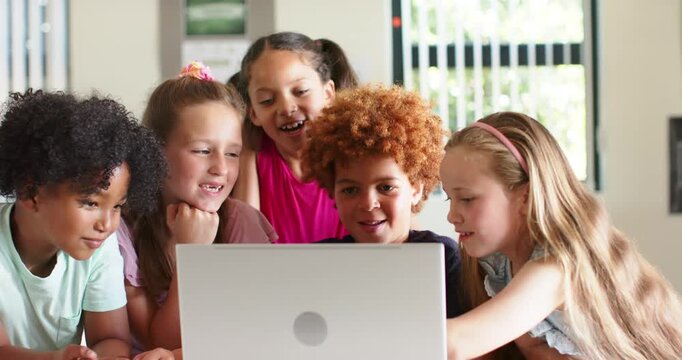 Diverse children friends leaning around laptop at classroom table, reacting to clip and laughing