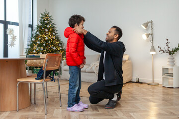 African American man helps his son put on his down jacket in the winter, father takes care of the child and gets him ready for school