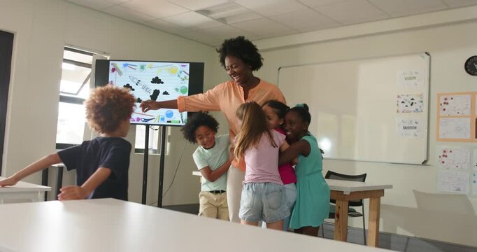 Teacher and students in classroom pointing at screen, child running in, children hugging for hug