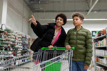African American woman and her son are shopping for groceries at supermarket, teenager and his mother are walking with cart through the store and smiling