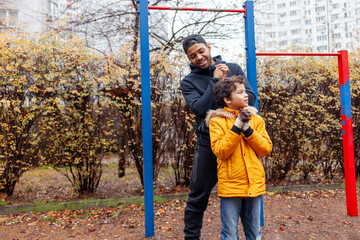 African American man and his son exercise and do exercises outdoors on playground, teenager stretches with his father on exercise equipment