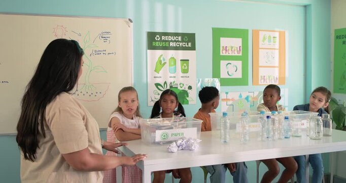 Diverse class teacher asking question and explaining recycling using bin labeled PAPER and bottles