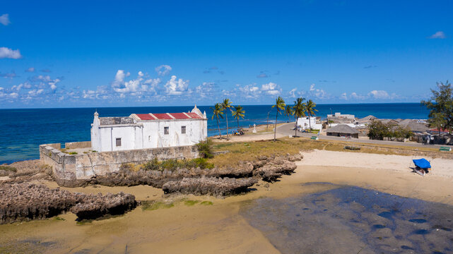 Aerial view of the Chapel of Nossa Senhora de Baluarte's weathered white facade against the turquoise sea, the coral rocks leading to the sandy beach, Ilha de Mocambique, Nampula, Mozambique.