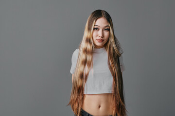 Woman with long hair wearing a white crop top standing against a gray background. Portrait of...