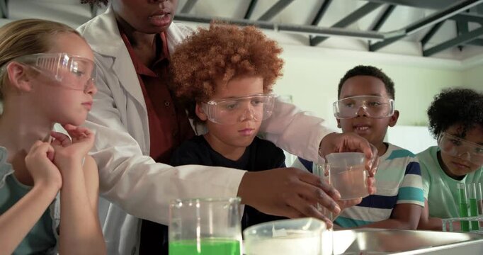 Female teacher teaching youth in lab, pointing, steadying as curly youth pouring cup into cylinder