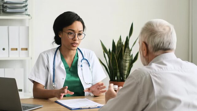 Female doctor in white lab coat and green scrubs explains a diagnosis to an attentive elderly male patient in a modern office with plant and medical tools. Concept of empathetic medical care