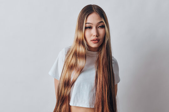Young woman with long straight hair standing against gray background wearing casual white t shirt looking at camera indoors with natural expression and posture.