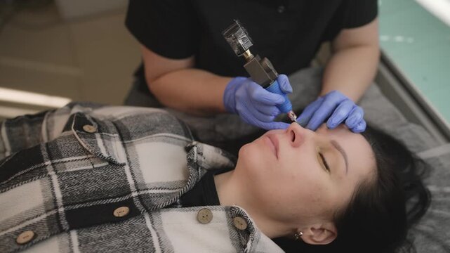 Close-up view of a beautician applying permanent makeup to a woman's eyebrows. The esthetician uses a special tool for the microblading procedure