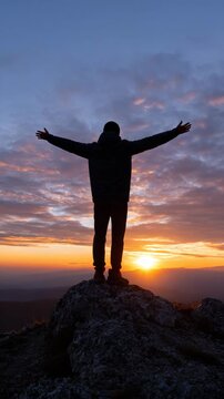 A person stands on a rock with arms outstretched at sunset.