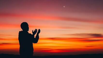 Fototapeta premium Silhouette of a person praying at sunset with vibrant orange and red clouds and a visible moon in the sky
