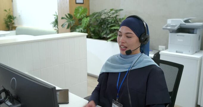 Woman with navy scarf at reception receiving call, answering headset, typing on keys helping caller