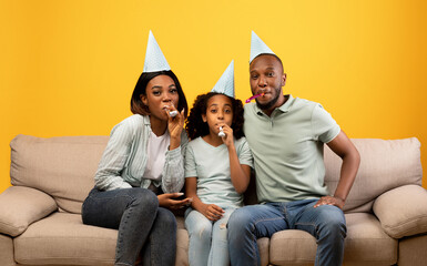 Happy african american parents and girl celebrating birthday in festive caps, looking at camera and blowing at party horns while sitting on sofa over yellow background