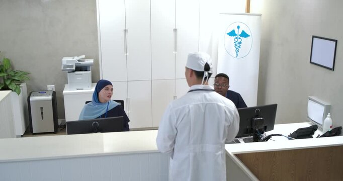 Asian man clinician approaching desk monitors, speaking and gesturing to clarify patient schedule