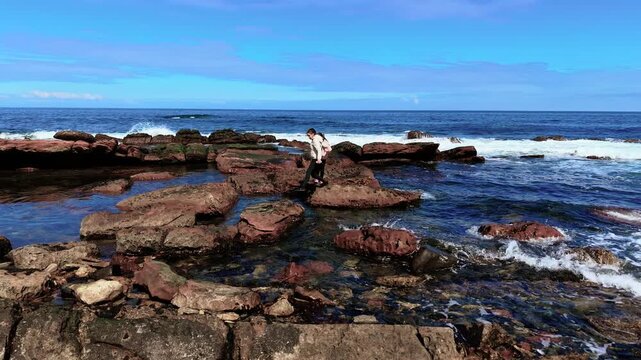 Mother and daughter jumping across rocks on sea shore. Family leaping over stones by ocean during coastal walk. Woman and girl playing near waves and skipping on seaside boulders
