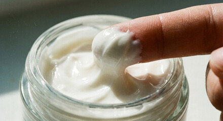Extreme macro of finger scooping thick luxury white face cream from a glass jar, tactile cosmetic texture.