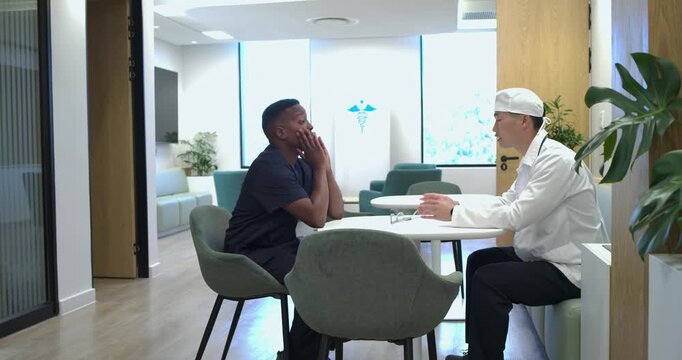 African-American patient upset covering face listening as Asian Dr reassuring with steth in clinic