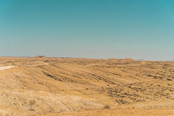 Naklejka premium On the way to Swakopmund, an endless road stretches through the striking landscape of the Namib Desert. Vast open plains, arid terrain, and gentle sand dunes meet a wide, dramatic sky.