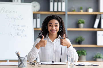 Excellent study result, positive emotions and distance education. Happy glad millennial cute african american female in headphones on blackboard background looking at webcam and showing thumbs up
