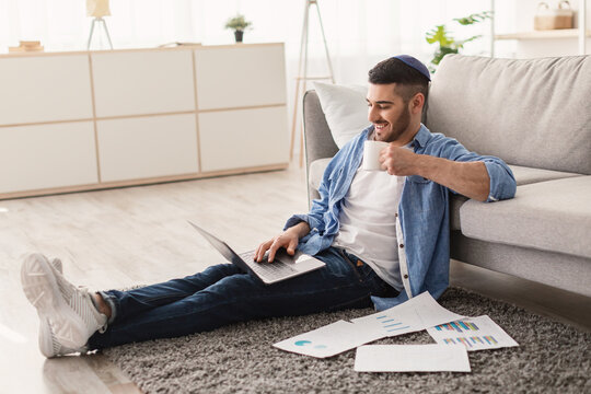 People And Technology Concept. Portrait of smiling young jewish man in kipa using laptop holding cup and drinking hot coffee. Male sitting on floor carpet with paperwork, watching video, taking break