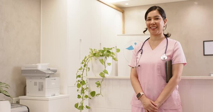 Female nurse standing right clinic desk with clipboard and stethoscope, greeting as camera zooming