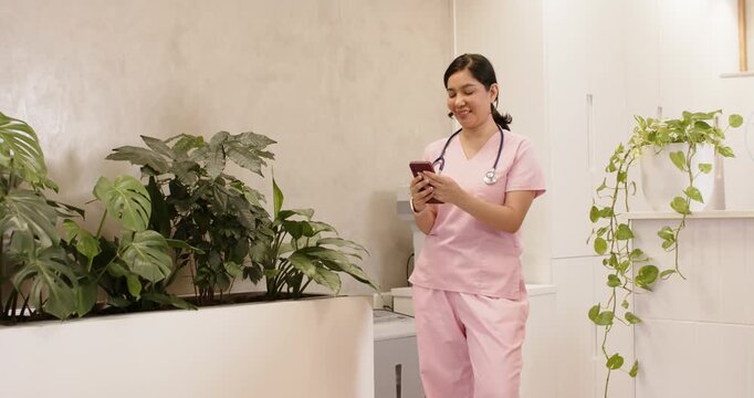 Adult female nurse in pink scrubs with stethoscope at clinic using phone reacting to alert smiling