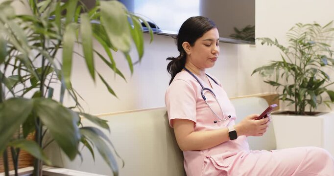 Adult woman in pink scrubs sitting on clinic bench, using phone, stethoscope, smiling after alert