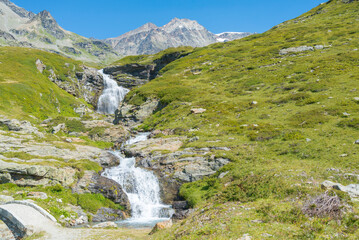 Mountain creek coming from the glaciers of the peaks of Italian Alps, Aosta Valley, Italy. Green meadow on the left. Blue sky on the background. With copy-space.