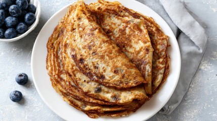 Golden folded pancakes arranged on white plate with scattered blueberries and neutral fabric. Top view of homemade breakfast or brunch, rustic food, sweet meal and comfort food concept