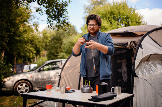 A man stands beside a tent, connecting parts of a camping stove on a table with outdoor cooking gear and a kettle.