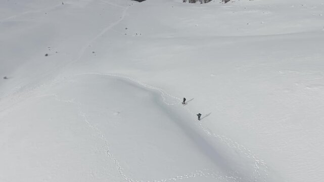 Aerial wide shot of two women hiking with a Czechoslovakian Wolfdog across a vast snowy mountain slope. Drone perspective of friends trekking in Abruzzo, Italy.