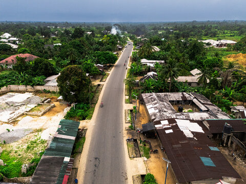 Aerial view of a long, straight road cutting through lush greenery and modest buildings under a hazy sky, Gokana, Rivers, Nigeria.