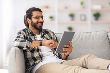 Relaxed young indian man chilling at home with digital tablet, sitting on couch, copy space. Happy guy freelancer shopping online, using modern pad and smiling, entertainment concept © Prostock-studio
