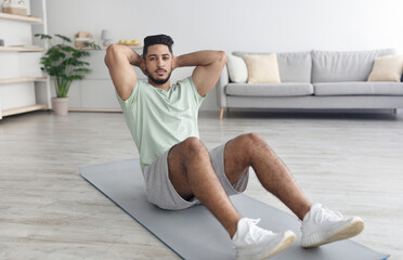 Obraz premium Sporty millennial Arab man doing abs exercises during domestic training, full length. Fit middle Eastern guy working out at home during covid lockdown, strengthening core muscles