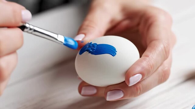 Person paints blue design on white egg with brush. Bright and clean setting at home with light wooden table. Concept of crafting, DIY projects, creativity