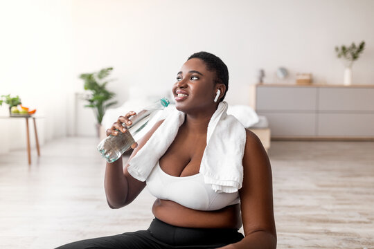 Portrait of young plus size black woman with earphones drinking water and listening to music on break from home training. Curvy African American lady staying hydrated after domestic workout