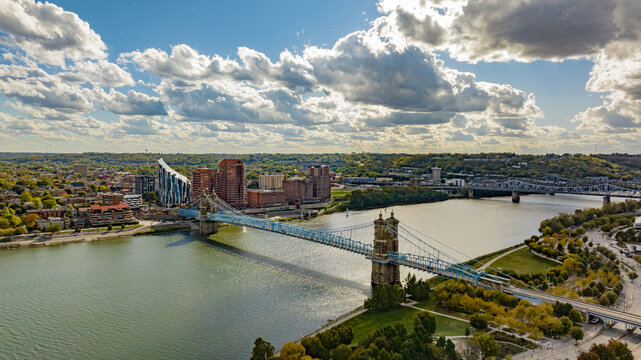 Aerial view of the majestic Roebling Suspension Bridge arches over the serene river, framed by autumnal foliage and cityscapes under a vast sky, Covington, Kentucky, United States.