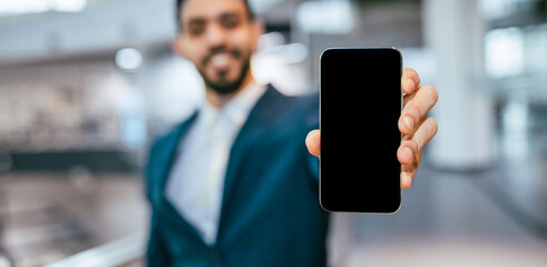 A man dressed in a suit is smiling and holding a smartphone out in front of him. The scene is in a...