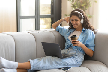 Happy young brunette woman in headphones having online video conference on laptop, drinking coffee...