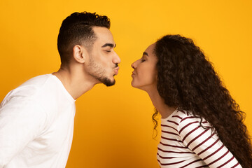 Side View Shot Of Cheerful Young Arab Couple Reaching Each Other With Lips, Ready To Kiss. Romantic Middle Eastern Man And Woman Expressing Love And Affection Over Yellow Studio Background, Closeup