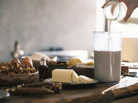 A hand pours creamy milk into a glass while surrounded by fresh butter, eggs, and grated lemon. The cozy kitchen exudes warmth and a homely atmosphere, perfect for baking