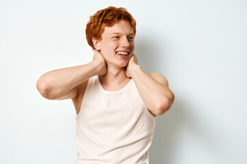 Young man with red hair wearing white sleeveless shirt smiling and looking away with hands on neck against plain white background in studio. Casual and happy expression.