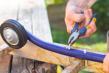 A man repairs a roof ladder wheel with pliers. The close-up photo shows hands working on the blue metal frame with the black wheel.