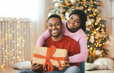 Obraz premium A couple sits close together on a soft surface, smiling while holding a wrapped gift. Behind them is a decorated holiday tree with lights. The atmosphere feels festive and joyful.