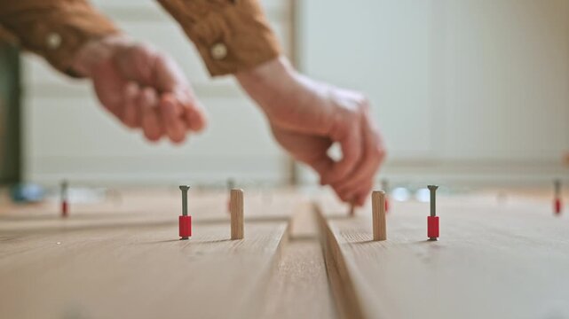 Hand of a skilled craftsman placing wooden dowels into pre-drilled holes on a wooden surface, demonstrating precise assembly techniques in a well-lit workspace