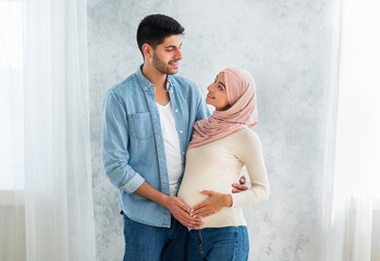 Fototapeta premium Happy parents-to-be. Happy muslim couple waiting for baby, man embracing wife and touching tummy, standing at home interior, free space. Islamic spouses cuddling and enjoying future parenting