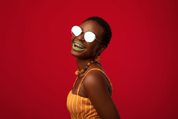 Emotional young black lady in summer outfit and stylish mirror sunglasses posing on red studio background, closeup profile portrait, copy space. Eyeglasses, eyewear, spectacles concept © Prostock-studio